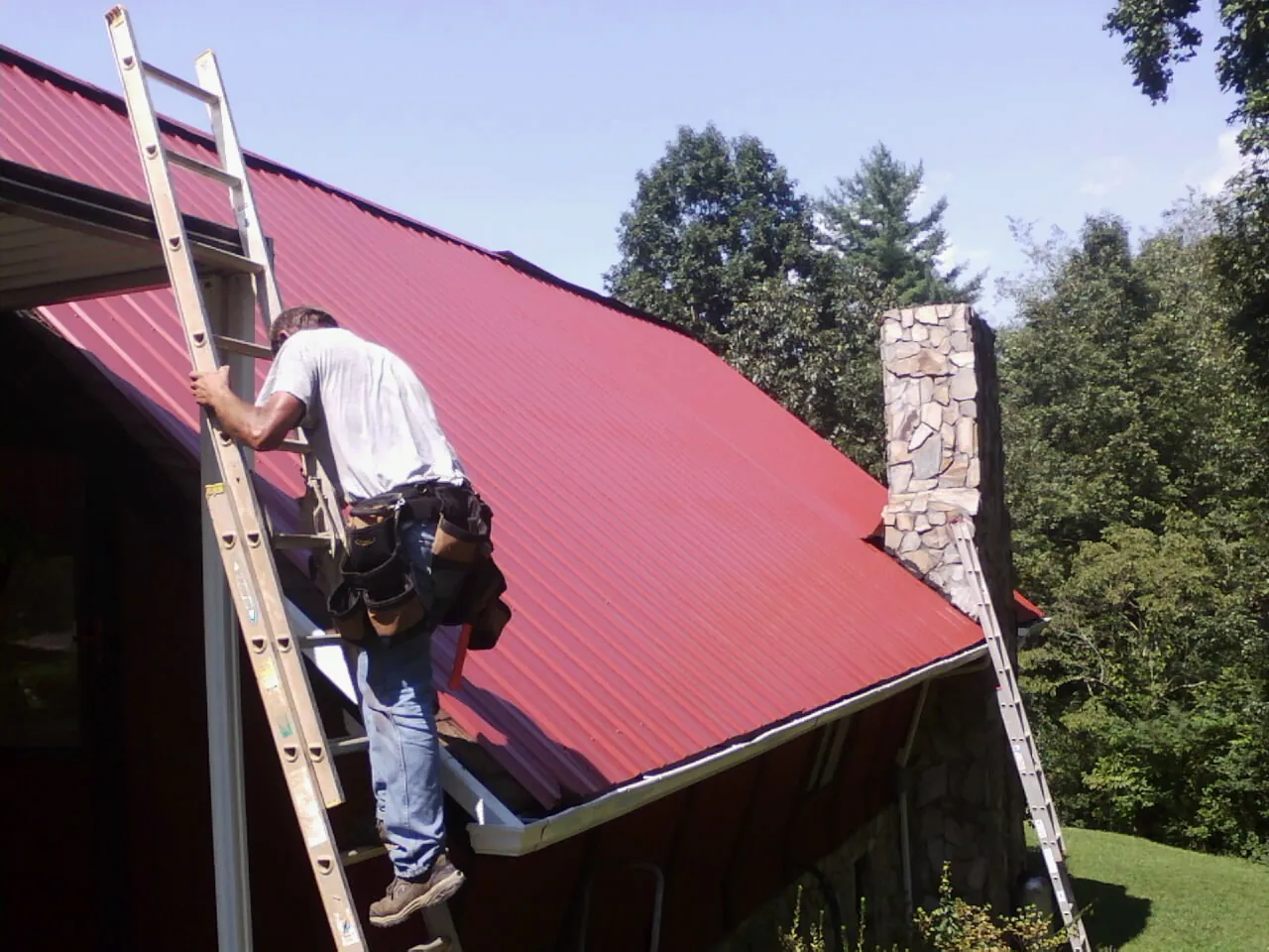 Roofer on a ladder beside a red metal roof and stone chimney — Soffit & Fascia Repair by Roofing Brothers in Taneytown